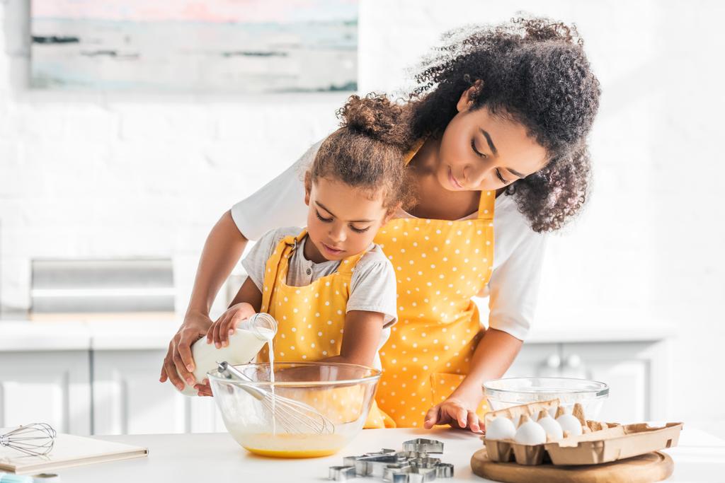 afro-americana mãe e filha preparando massa e derramando leite em tigela na cozinha
 - Foto, Imagem