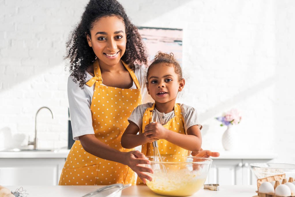усміхнена афроамериканська дочка і мати готують і збивають тісто на кухні, дивлячись на камеру
 - Фото, зображення