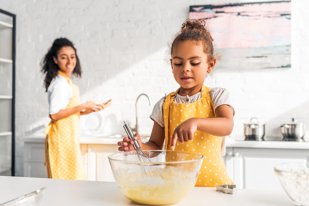 hija afroamericana preparando y batiendo la masa, poniendo el dedo en un tazón en la cocina
 - Foto, imagen