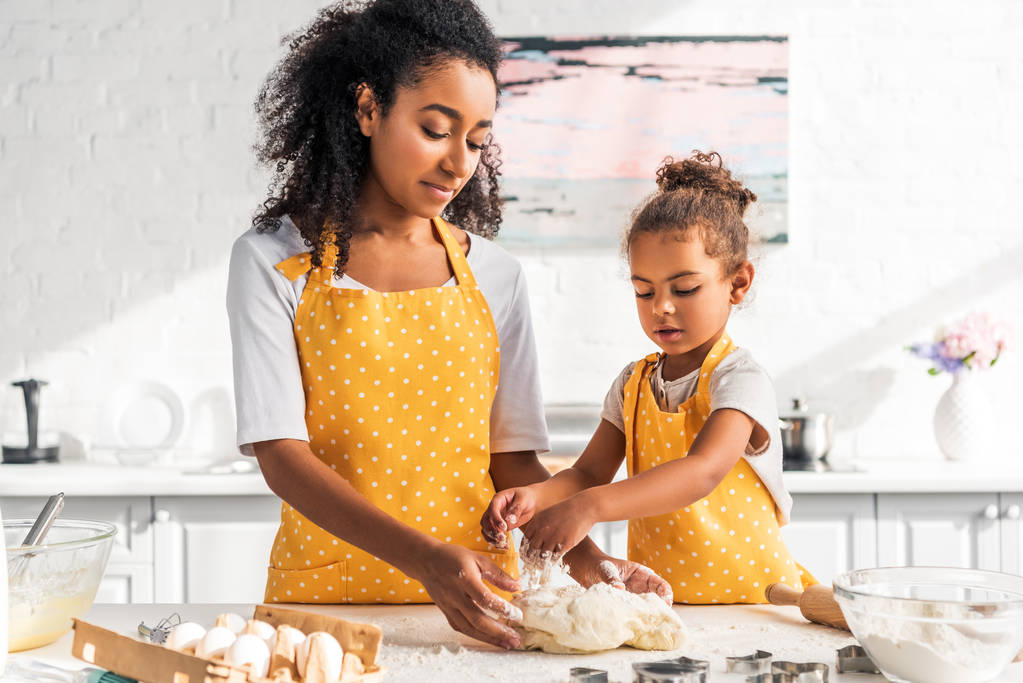 Afro-Amerikaanse moeder en dochter in schorten kneden van deeg met de handen in de keuken - Foto, afbeelding
