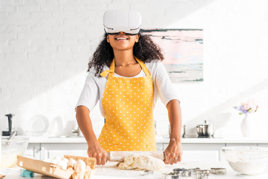 χαμογελώντας αφρικανικό αμερικανικό κορίτσι στην ποδιά και σετ κεφαλής εικονικής πραγματικότητας κυλώντας ζύμη στην κουζίνα - Φωτογραφία, εικόνα