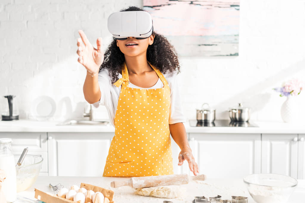 podekscytowany african american girl w fartuch i zestaw wirtualnej rzeczywistości, osiągając sił w tabeli z ciasta w kuchni - Zdjęcie, obraz