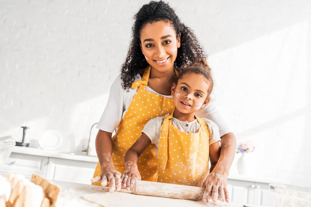 vrolijke Afro-Amerikaanse moeder helpt dochter deeg met de deegroller rollen in keuken, camera kijken - Foto, afbeelding