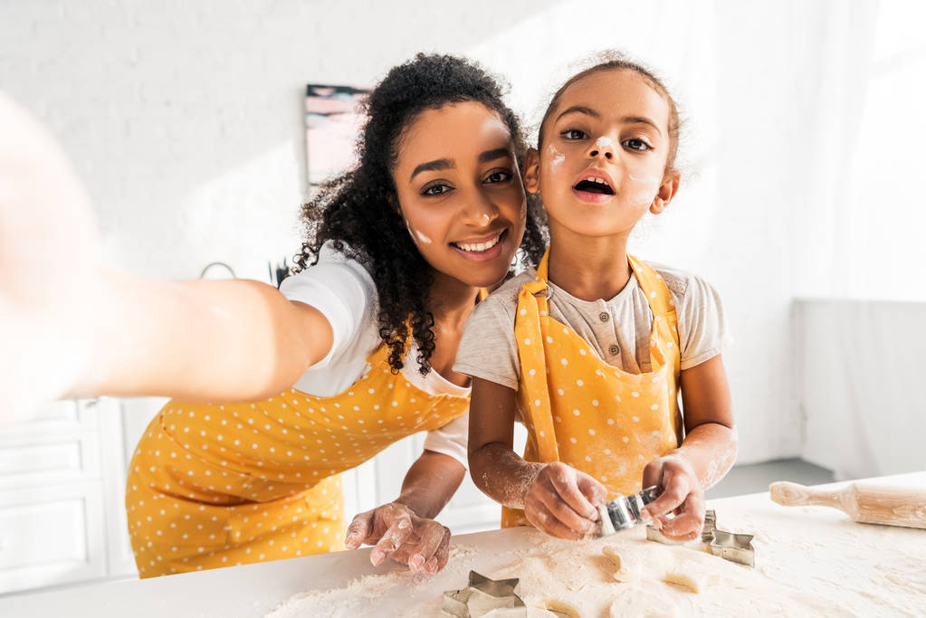 ponto de vista da câmera de sorrir afro-americana mãe e filha em aventais preparar biscoitos na cozinha
 - Foto, Imagem