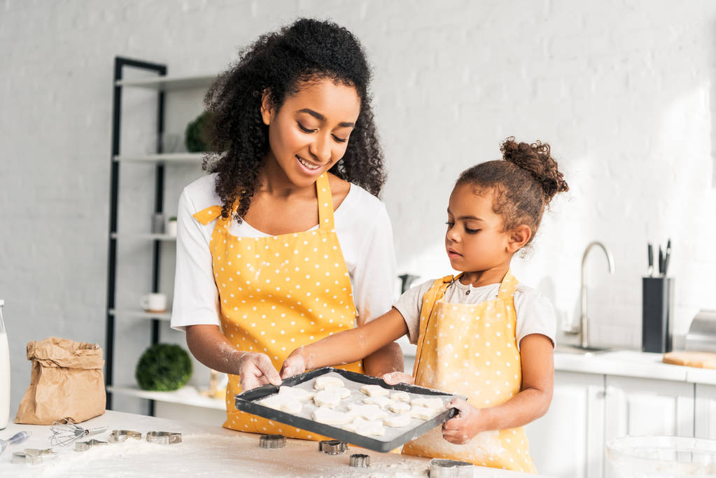 Αφρικανική αμερικανική μητέρα και κόρη εκμετάλλευση δίσκος με unbaked μπισκότα μαζί στην κουζίνα - Φωτογραφία, εικόνα