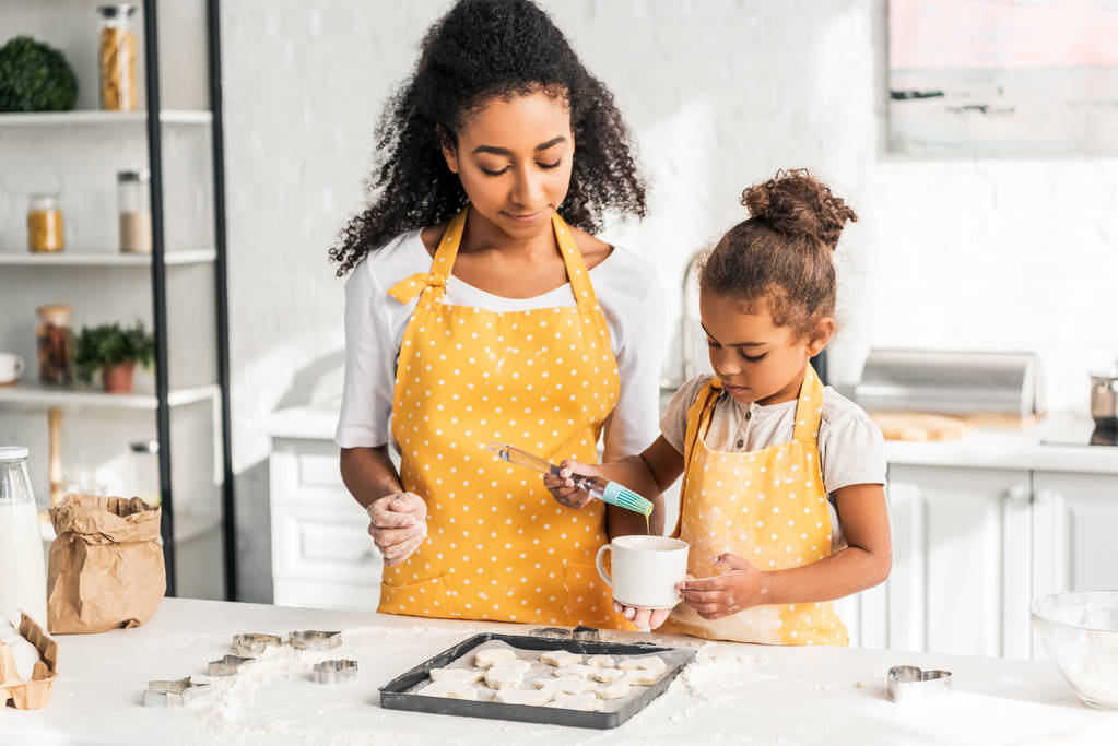 Afro-Amerikaanse moeder en dochter olie aanbrengen op de ongebakken koekjes in keuken - Foto, afbeelding