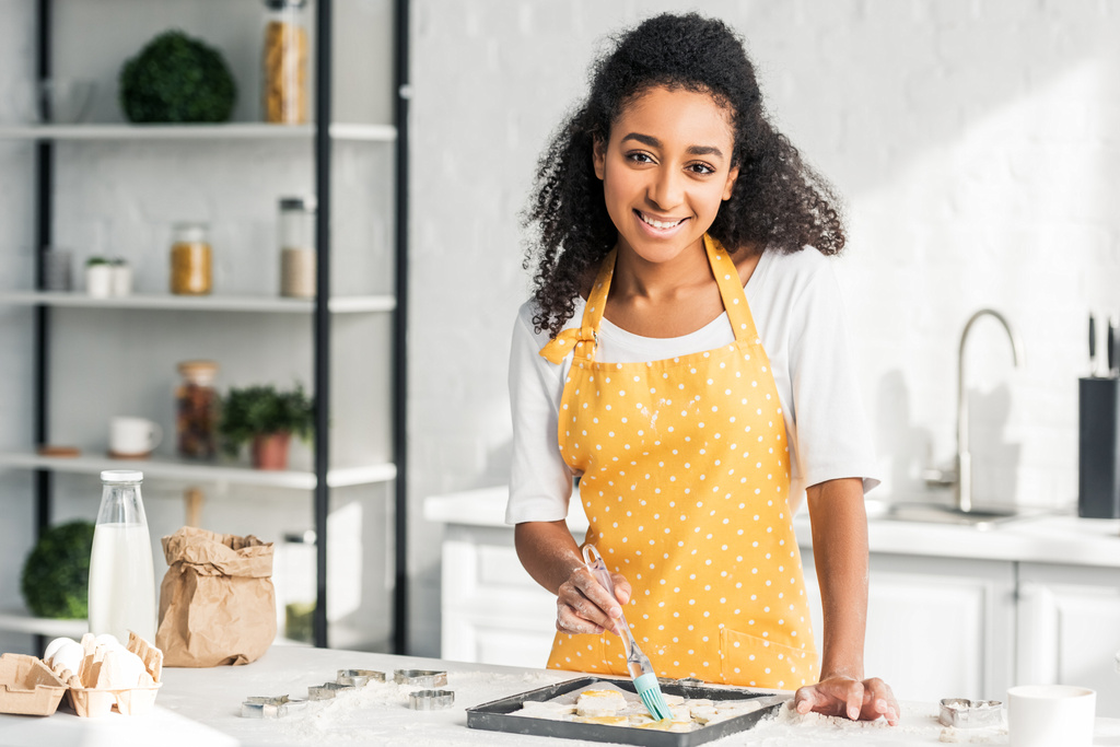 schöne lächelnde afrikanisch-amerikanische Mädchen in Schürze Öl auf ungebackene Kekse in der Küche auftragen und in die Kamera schauen - Foto, Bild