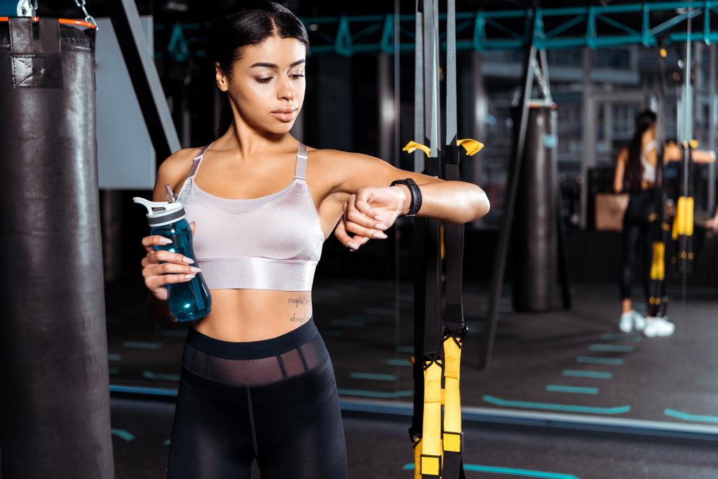 fantastisches sportliches Mädchen mit Wasserflasche beim Anblick des Fitness-Trackers in der Sporthalle  - Foto, Bild