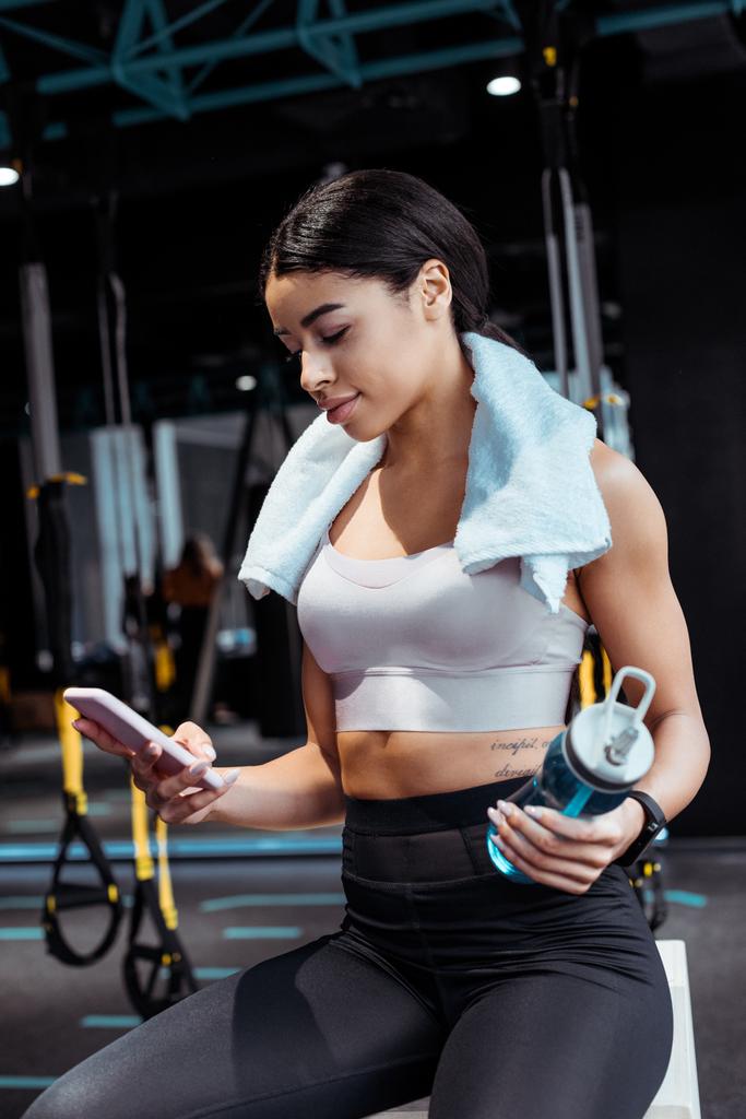 Hermosa chica deportiva usando dispositivo digital mientras sostiene la botella de deporte en el gimnasio
  - Foto, Imagen