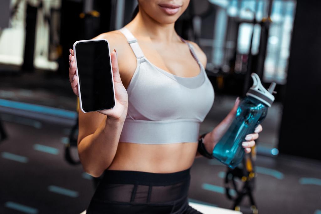 Przycięte widzenia sportive kobieta trzymając butelkę smartphone i sport w siłowni fitness  - Zdjęcie, obraz