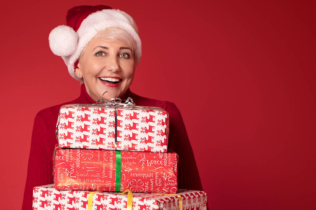 Happy smiling mature woman in red santa claus hat holding stack of gift boxes, on red studio background. Christmas shopping, copy space. - Photo, Image
