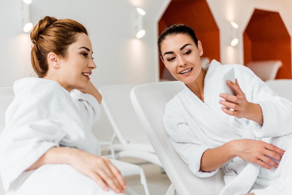 souriantes jeunes femmes regardant smartphone tout en se reposant ensemble sur des chaises longues dans le spa
  - Photo, image