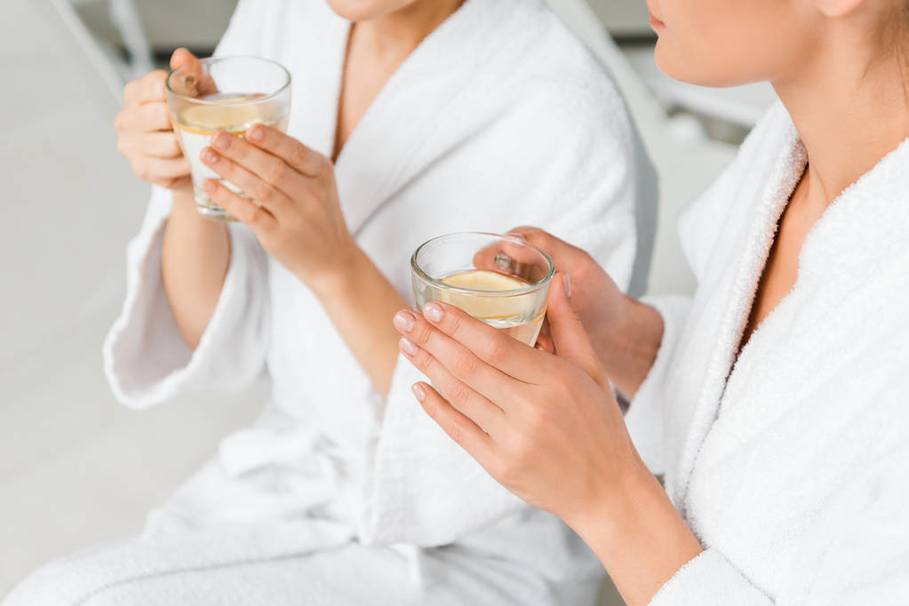 abgeschnittene Aufnahme junger Frauen in Bademänteln, die Tassen mit Kräutertee und Zitrone halten, während sie sich im Wellnessbereich ausruhen - Foto, Bild