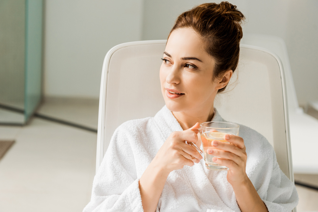 attraente giovane donna che tiene in mano una tazza di bevanda a base di erbe e distoglie lo sguardo mentre riposa sul lettino in spa
   - Foto, immagini