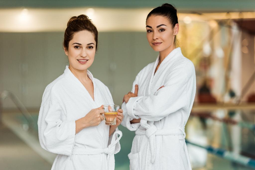 schöne Frauen in Bademänteln halten Tasse mit Kräutertee in der Hand und lächeln in die Kamera im Wellnessbereich - Foto, Bild