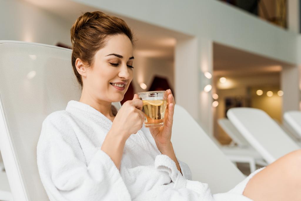 sonriente joven mujer sosteniendo taza con bebida de hierbas mientras se relaja en el spa
  - Foto, imagen
