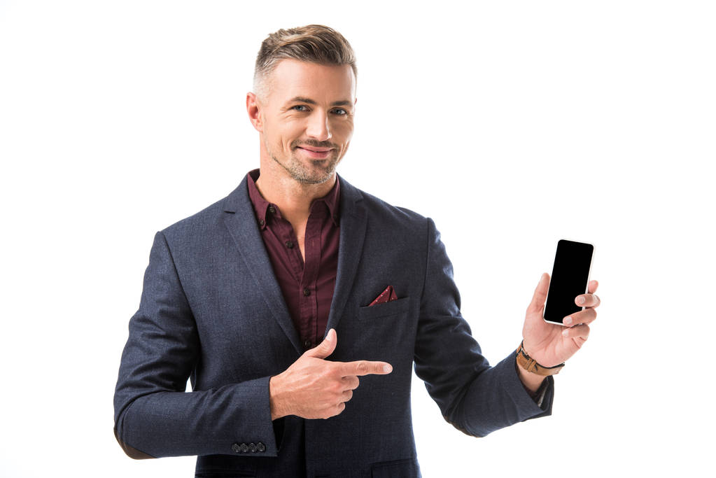 positive adult stylish man in jacket pointing by finger at smartphone with blank screen isolated on white - Photo, Image