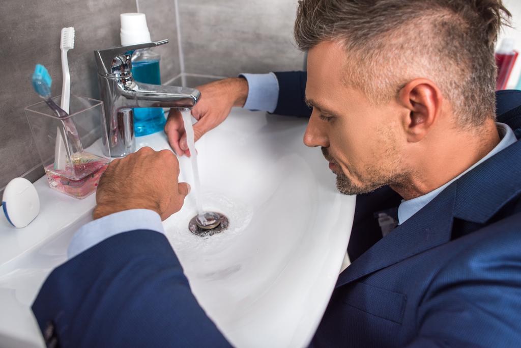 vista lateral del hombre de negocios adulto en traje sentado frente al lavabo en el baño
  - Foto, imagen