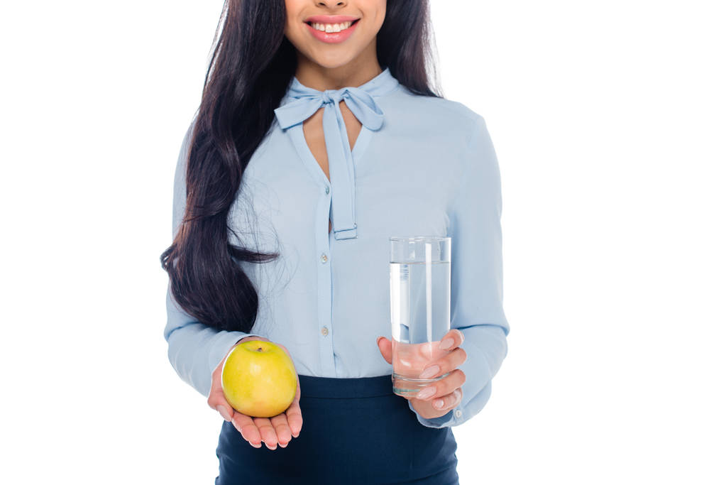 abgeschnittene Aufnahme einer lächelnden Afroamerikanerin mit einem Glas Wasser und einem Apfel auf weißem Grund - Foto, Bild