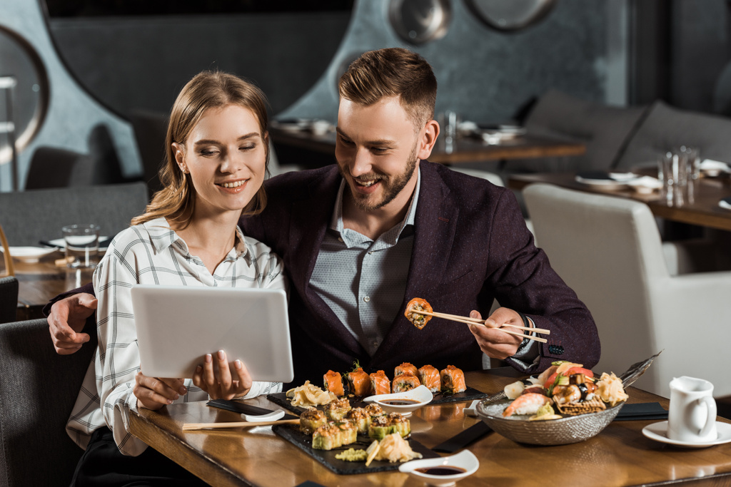 Atractiva pareja de yound adulto usando tableta digital mientras come sushi en el restaurante
 - Foto, Imagen