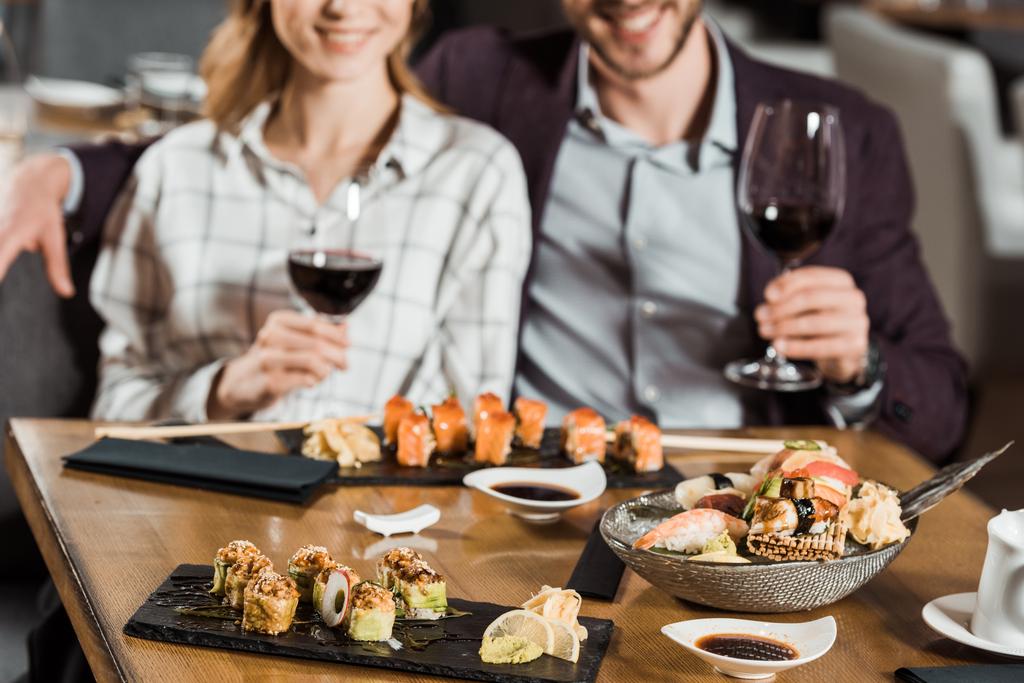 Частичный вид улыбающейся пары, которая ест суши и пьет вино в ресторане
 - Фото, изображение