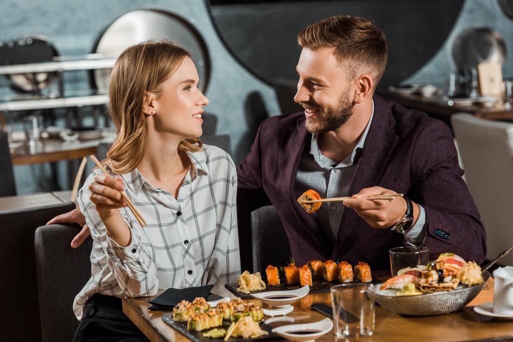 Boldog, fiatal felnőtt pár néztek egymásra, miközben étteremben enni sushi tekercs  - Fotó, kép