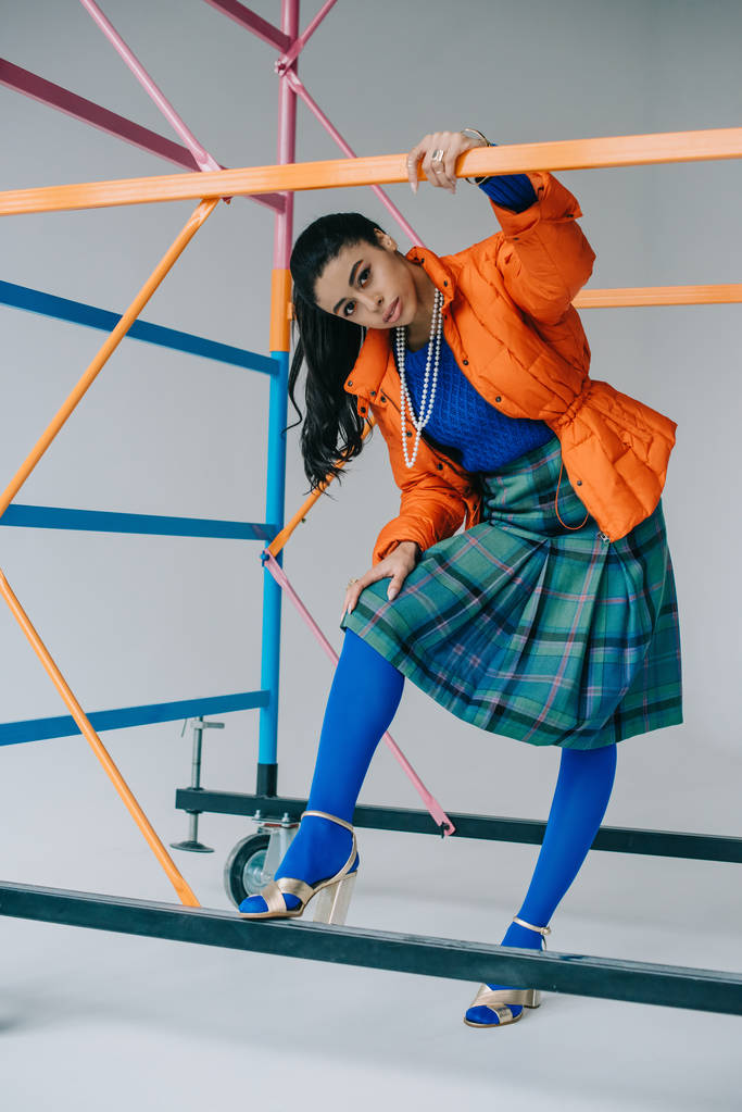 stylish girl in orange winter jacket, checkered skirt and blue tights posing near colorful scaffold in studio - Photo, Image