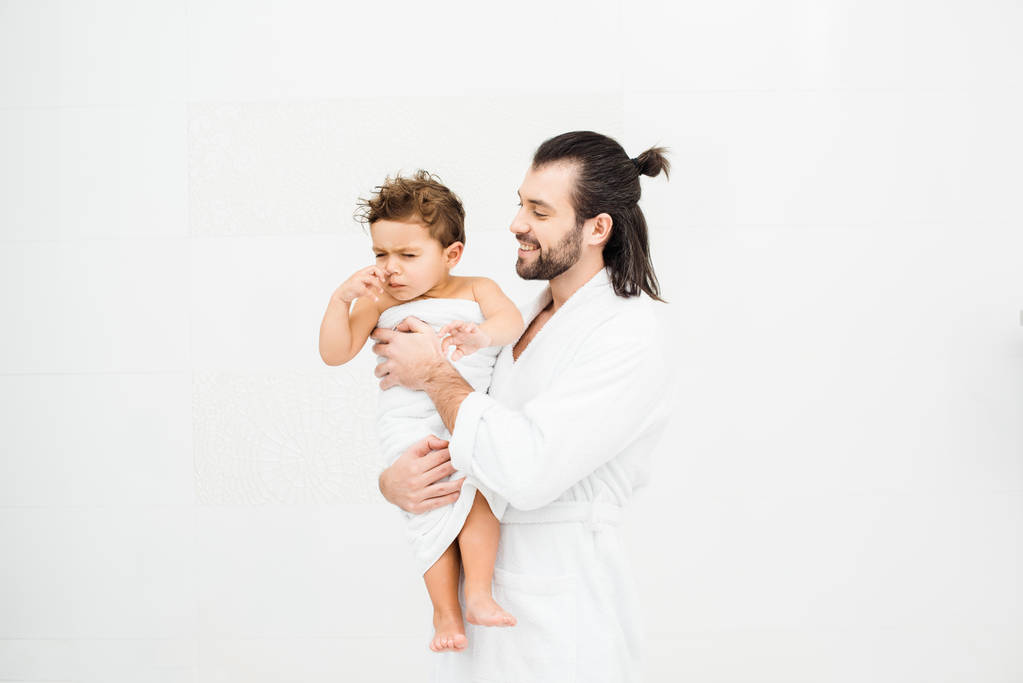 Father in bathrobe looking at son in towel and smiling on white - Photo, Image
