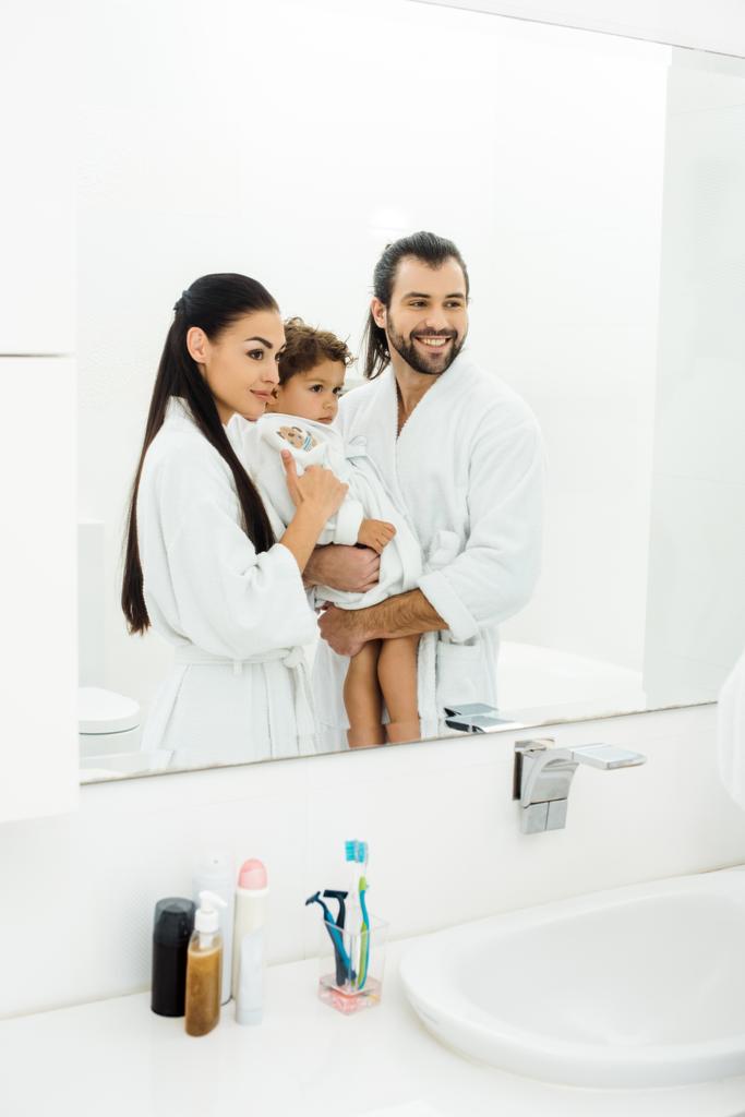 Семья смотрит в зеркало и улыбается в белой ванной комнате
  - Фото, изображение