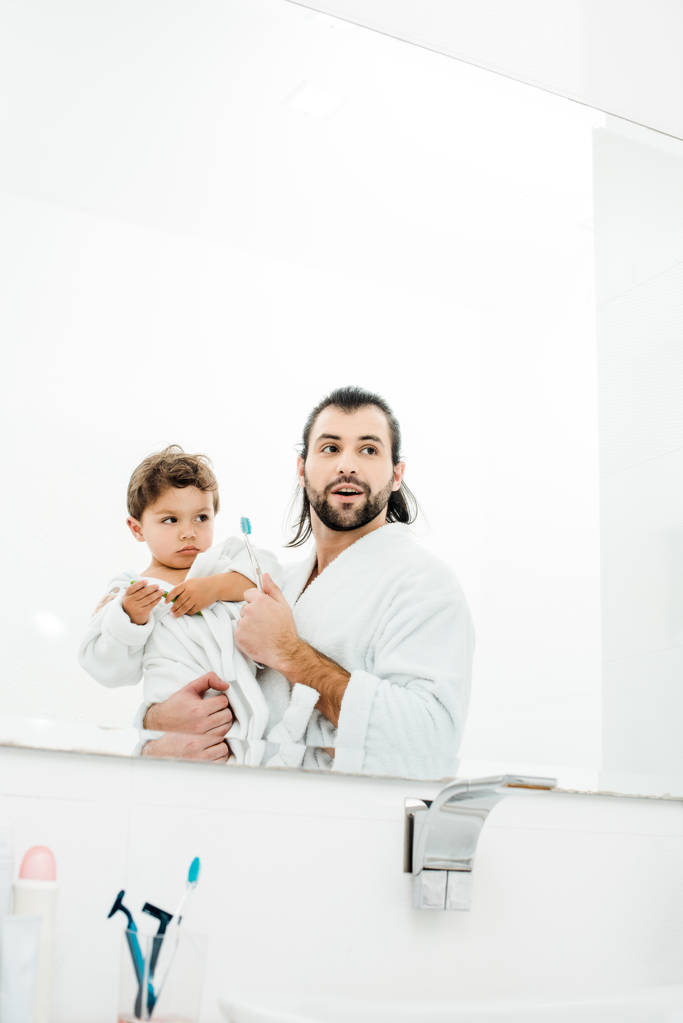 Μπαμπάς και γιος ψάχνουν στον καθρέφτη σε λευκή μπουρνούζια και κρατώντας οδοντόβουρτσες - Φωτογραφία, εικόνα