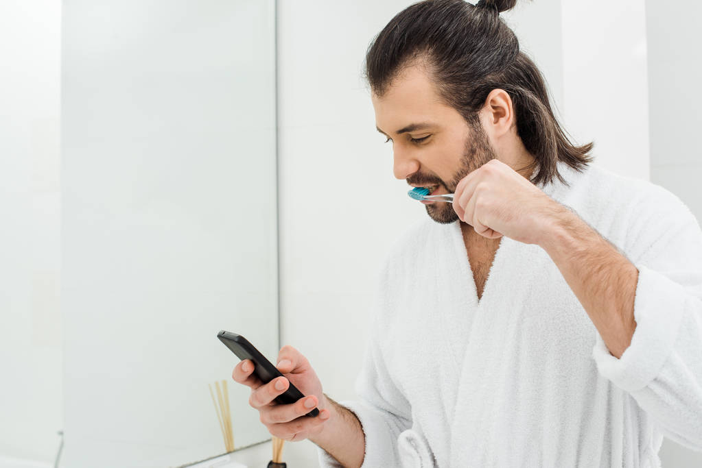 hombre adulto guapo usando teléfono inteligente y cepillarse los dientes en el baño
 - Foto, imagen