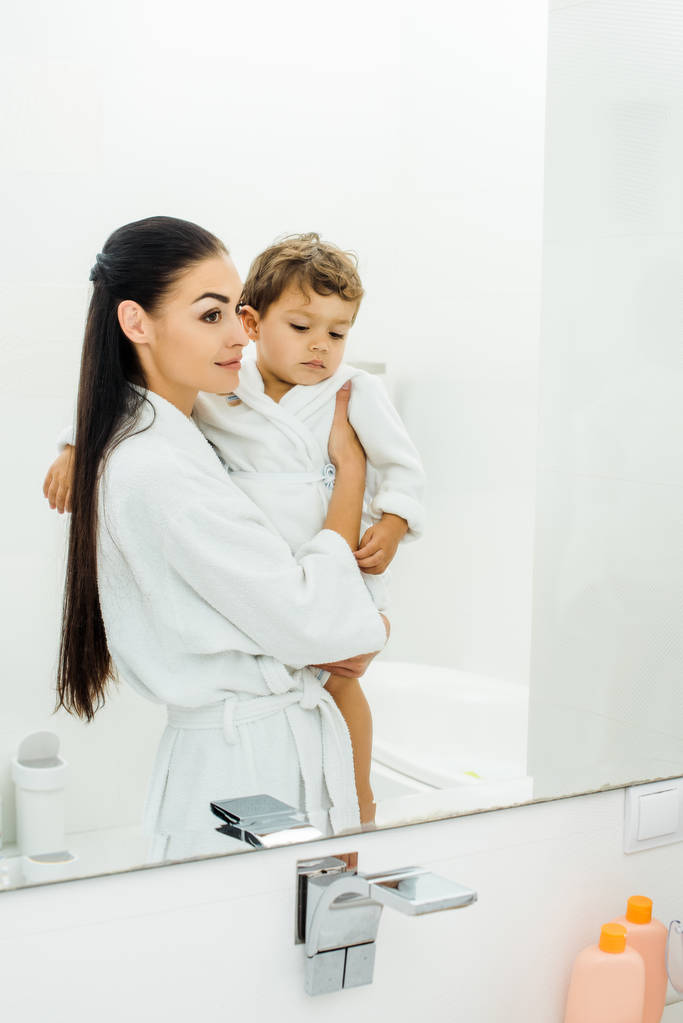 mooie moeder bedrijf zoon in witte badjas in badkamer - Foto, afbeelding