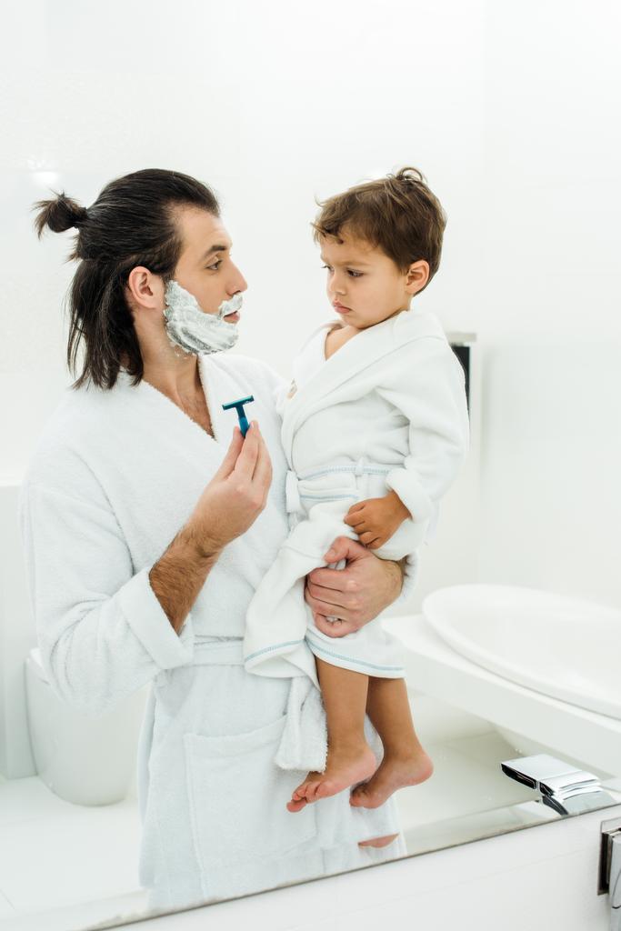 Isä valkoisessa kylpytakissa näyttää partaveitsi pikkulapselle poika
  - Valokuva, kuva