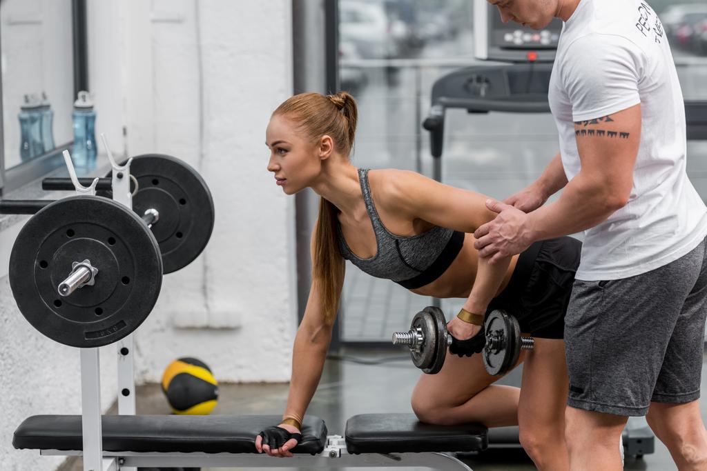 cropped image of personal trainer helping sportswoman exercising with dumbbells in gym - Photo, Image