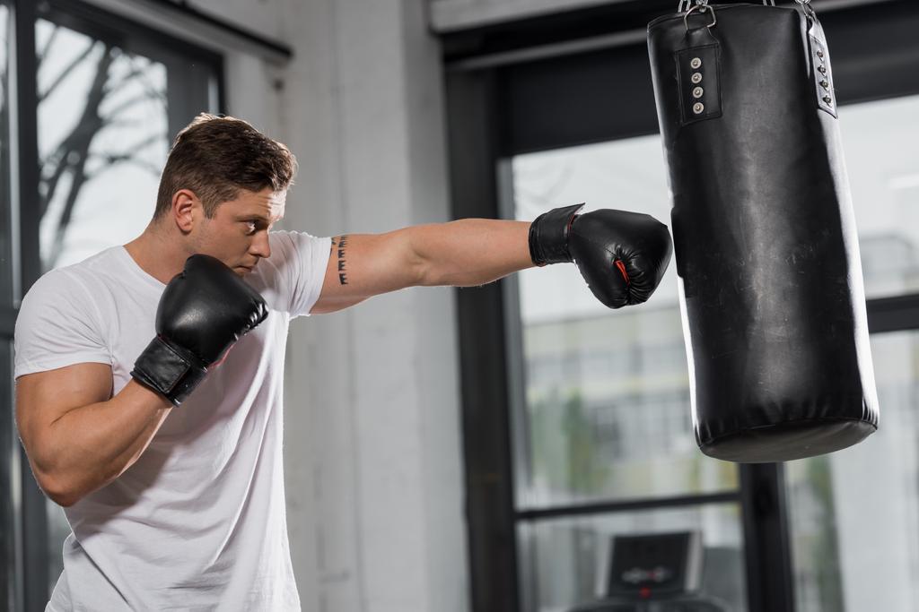 Zijaanzicht van knappe gespierde bokser traint met bokszak in sportschool - Foto, afbeelding