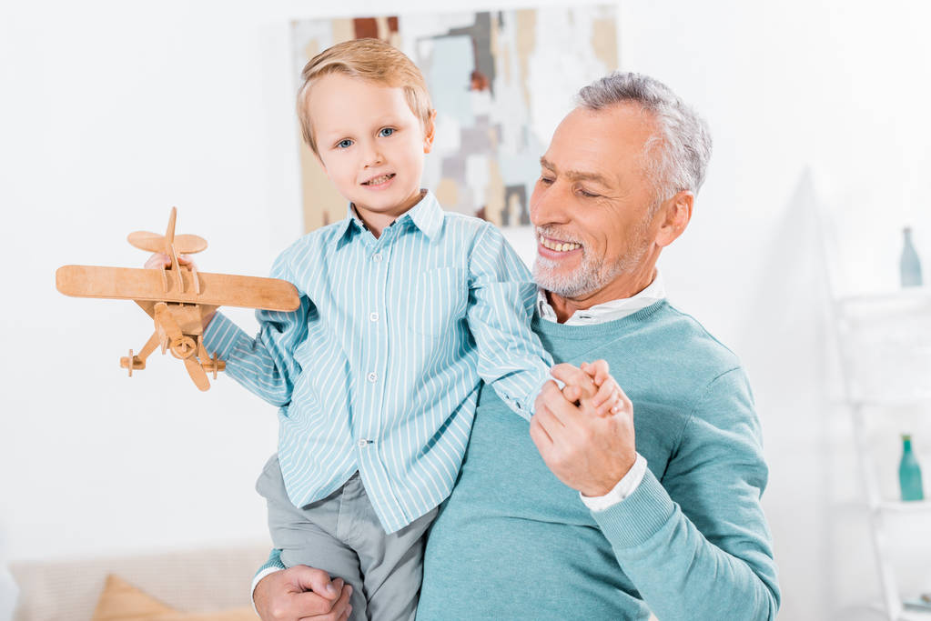 επιλεκτική εστίαση του χαμογελώντας μέσης ηλικίας άνδρα να κρατάει το μικρό εγγονό με ξύλινα αεροπλάνο στα χέρια στο σπίτι - Φωτογραφία, εικόνα