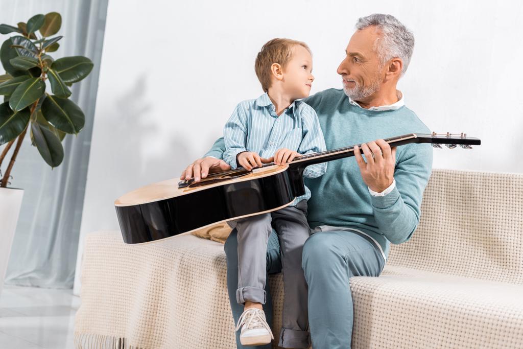 έκπληκτος ο παππούς με ακουστική κιθάρα βλέπουν αξιολάτρευτο μικρό εγγονό στον καναπέ στο σπίτι - Φωτογραφία, εικόνα