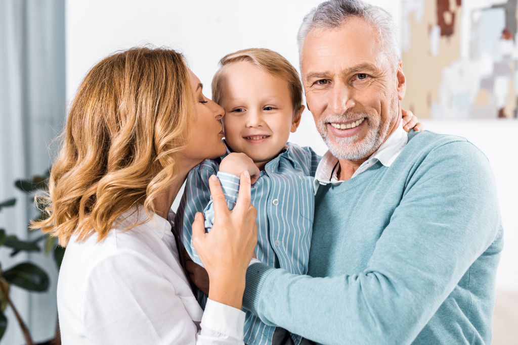 enfoque selectivo de alegre pareja de mediana edad sosteniendo nieto adorable y besándolo en las mejillas
 - Foto, Imagen