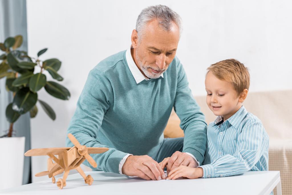 volwassen man die papier vliegtuig terwijl zijn schattige kleinzoon zitten in de buurt van aan tafel met houten vliegtuig  - Foto, afbeelding