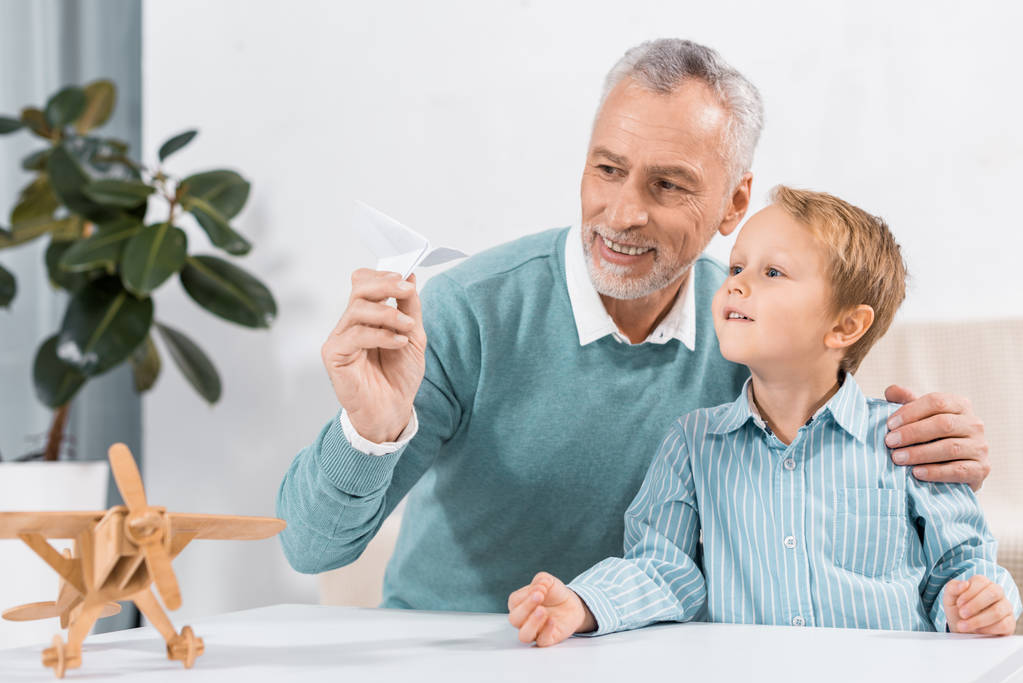 улыбающийся взрослый мужчина и внук веселятся с бумажным самолетом дома
 - Фото, изображение