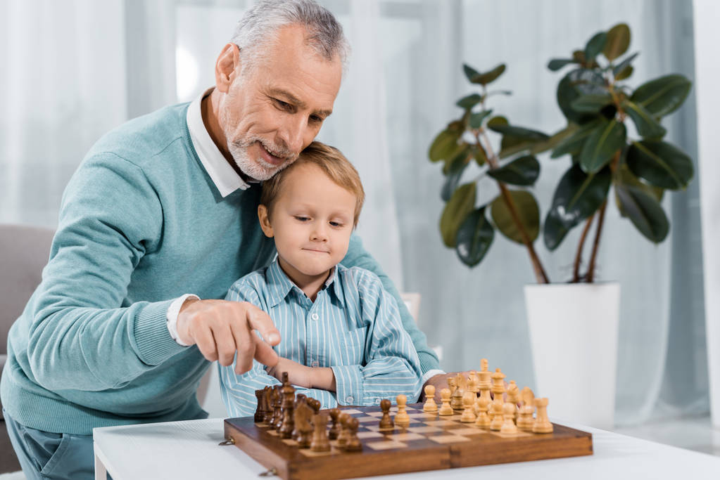 fröhlicher Mann mittleren Alters beim Schachspielen mit entzückendem Enkel zu Hause - Foto, Bild