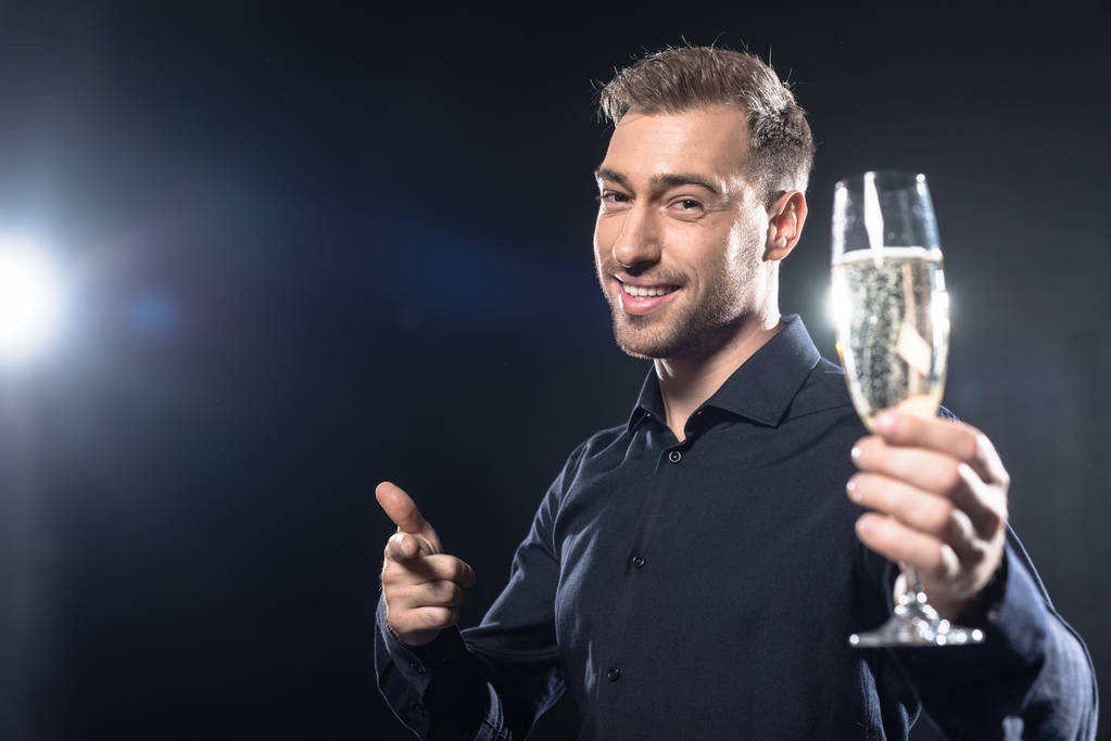 усміхнений молодий чоловік у стильному блістері тримає келих шампанського і вказує на камеру ізольовано на чорному
 - Фото, зображення