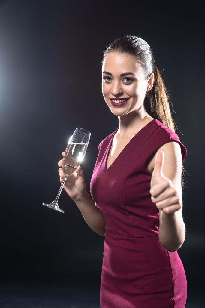 s úsměvem mladá žena drží sklenku šampaňského a objevil palec na kameru na černém pozadí - Fotografie, Obrázek
