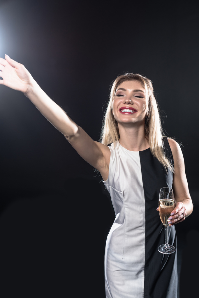 happy young woman holding glass of champagne and waving with hand on black - Photo, Image