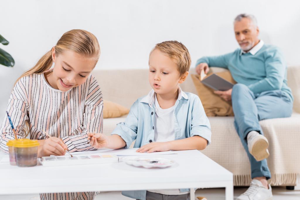 entzückende Kinder malen am Tisch, während ihr Großvater zu Hause auf dem Sofa ein Buch liest - Foto, Bild