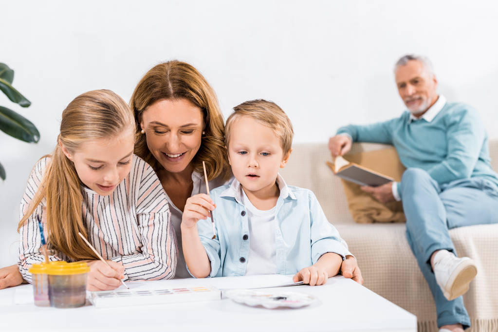 rijpe vrouw op zoek hoe haar kleinkinderen schilderen terwijl haar echtgenoot zitten achter op Bank bij home - Foto, afbeelding