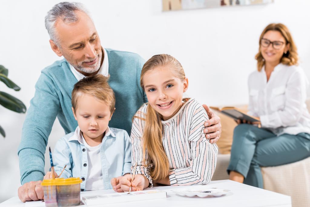 knappe middelste leeftijd man op zoek hoe kleinkinderen schilderen terwijl zijn vrouw zitten achter op home - Foto, afbeelding