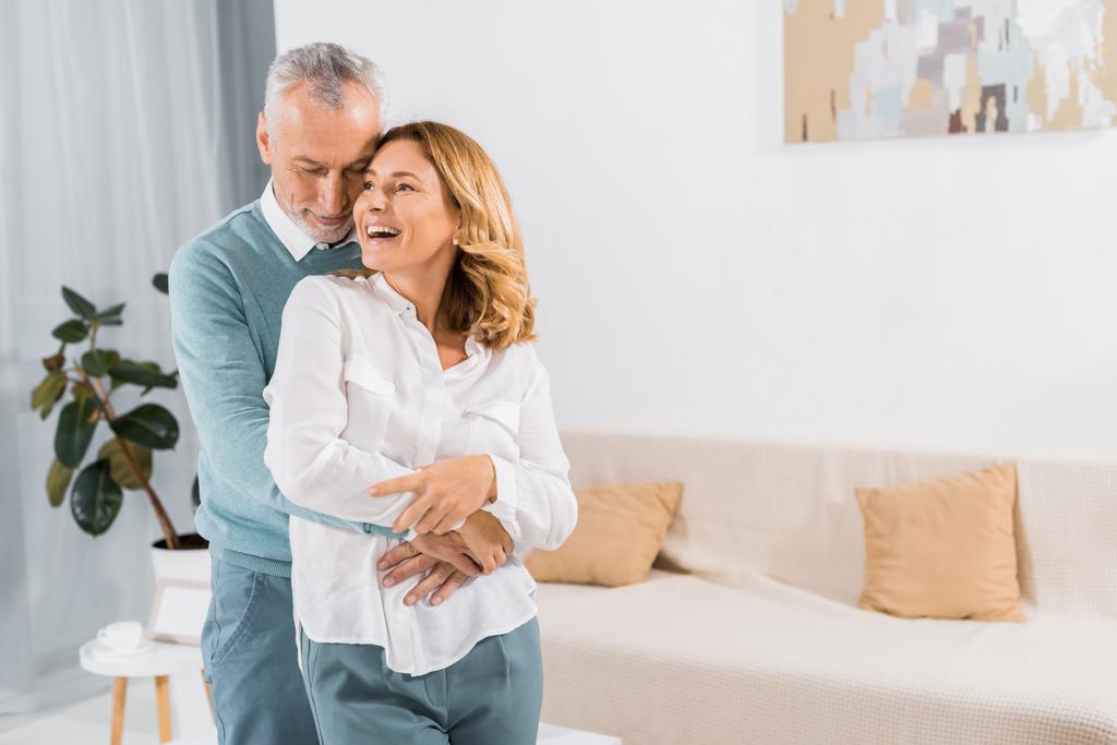 Mann mittleren Alters umarmt lachende Frau im heimischen Wohnzimmer - Foto, Bild