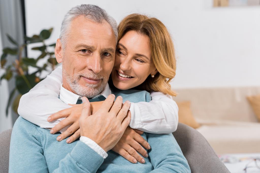 selektiver Fokus einer glücklichen attraktiven Frau, die ihren Mann mittleren Alters zu Hause umarmt - Foto, Bild