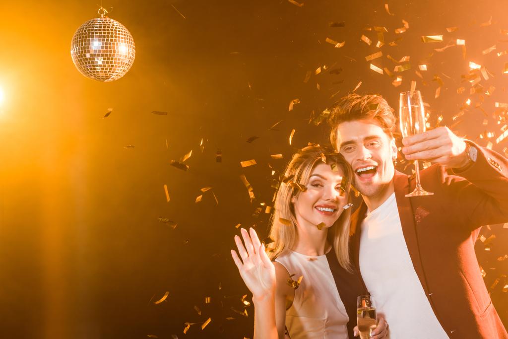glücklich flirtendes Paar mit Champagner beim Feiern unter goldenem Licht, während von oben Konfetti verschüttet wird - Foto, Bild